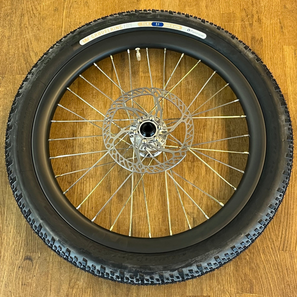 A black mountain bike wheel with a silver multi-spoke disc brake rotor, photographed from directly above on a light brown wooden floor.