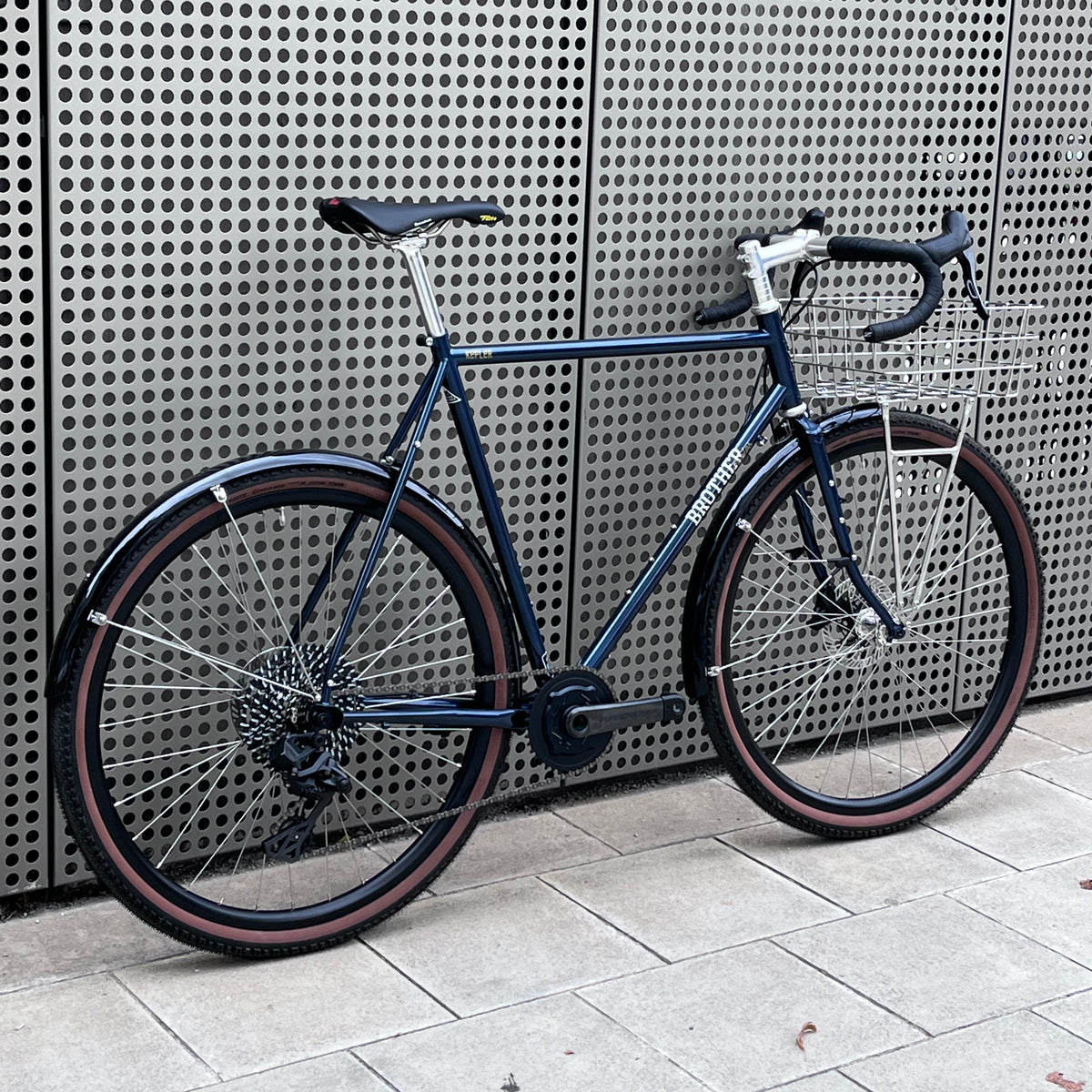 A dark navy blue vintage-style road bicycle with a rear cargo rack, leaning against a perforated metal wall on a light gray tiled floor.