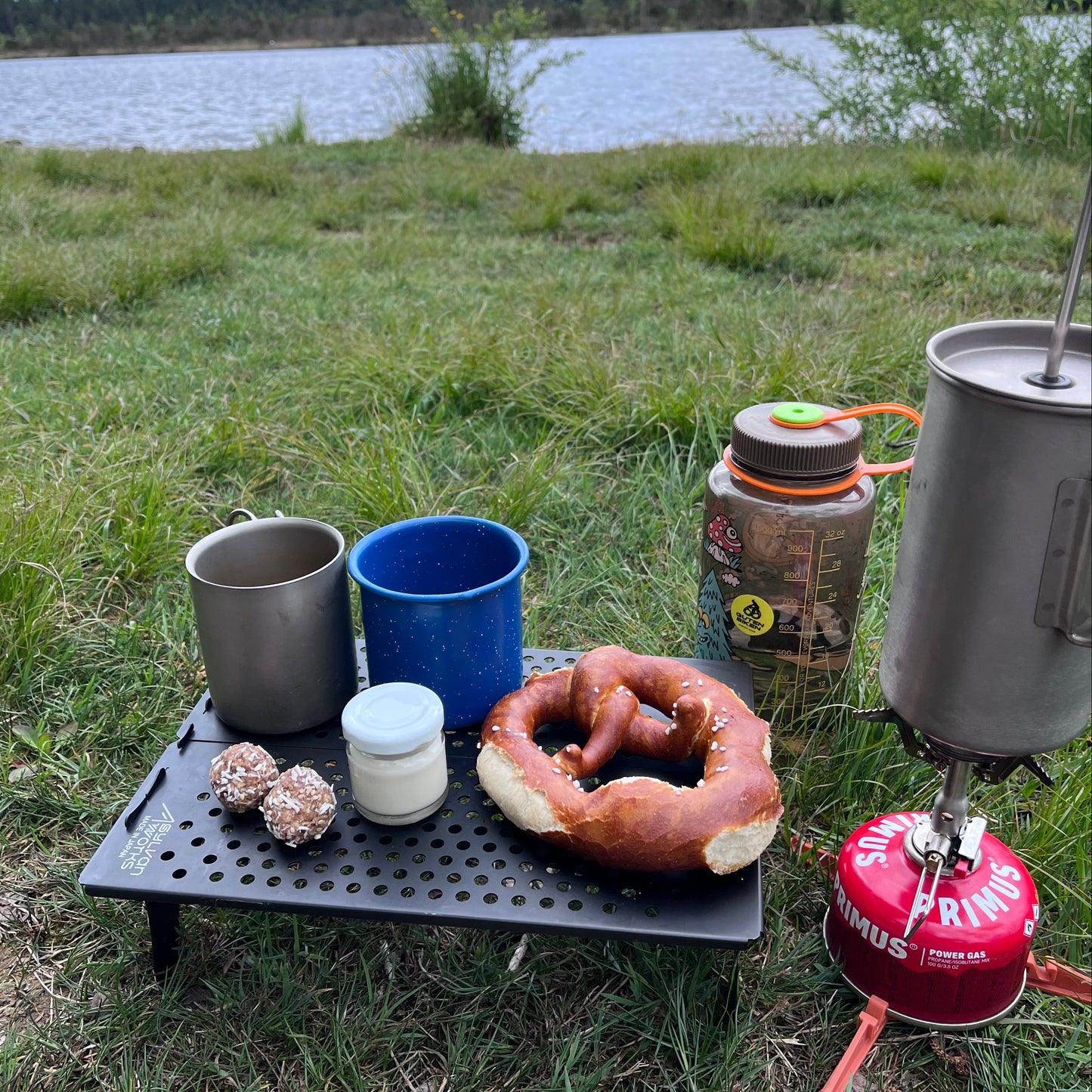 Camping setup with pots, cups, and a pretzel near a lake.