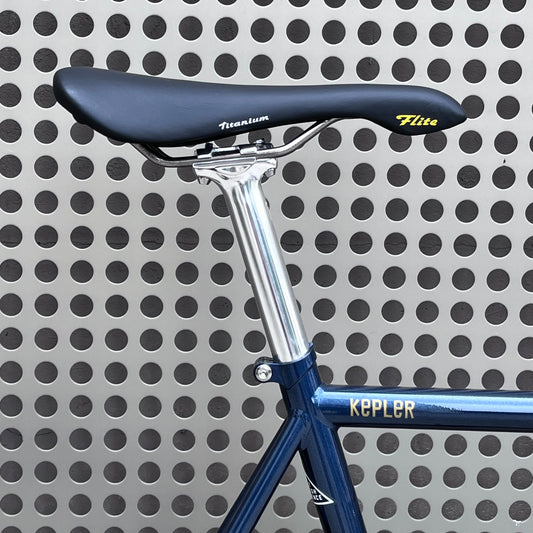Close-up of a bicycle saddle and seatpost with a blue frame, set against a perforated metal wall. The saddle is black with yellow and white branding, and the frame has 'KEPLER' in gold lettering.