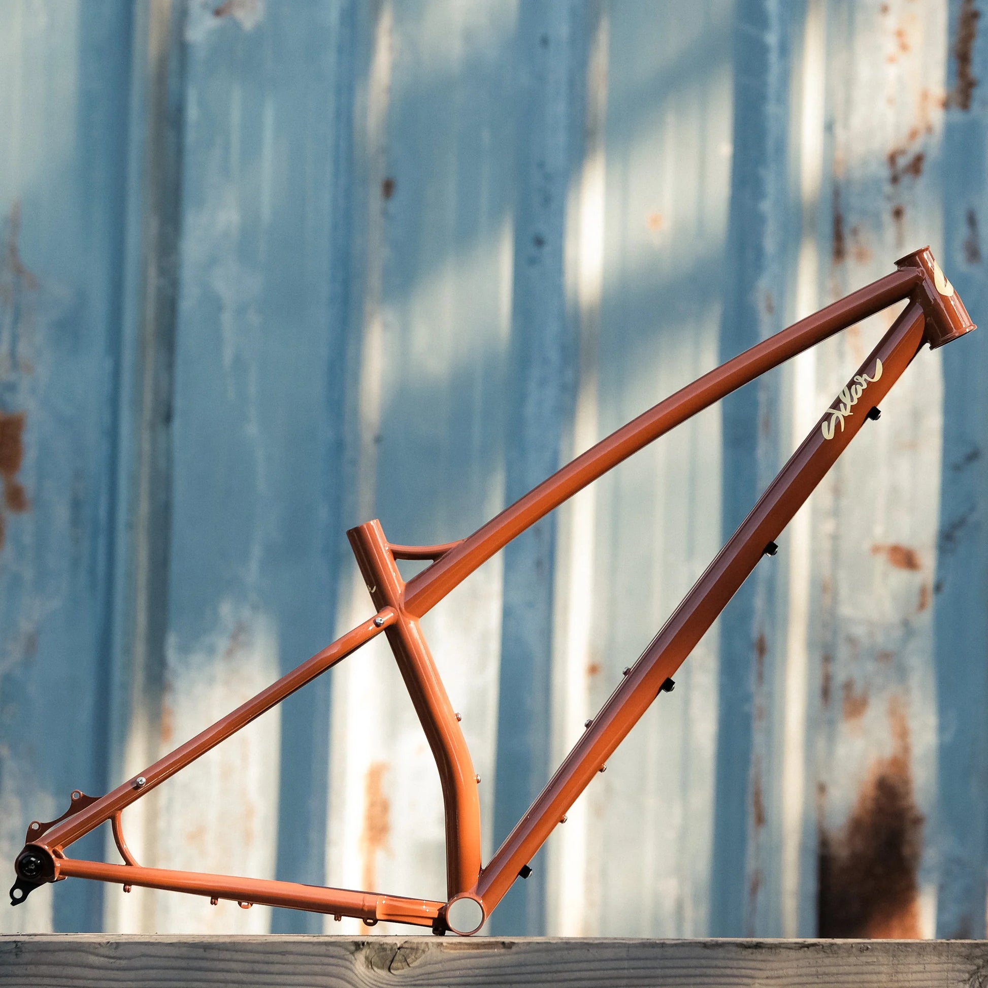 A orange hardtail mountain bike frame leaning against a rustic metal wall.